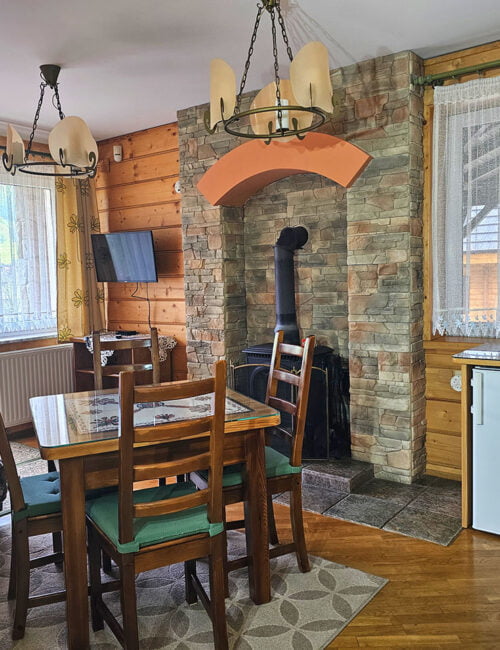 Salon utrzymany w stylu góralskim z drewnianym stołem, kanapą, telewizorem, kominkiem, parkietem, lodówka oraz boazerią na ścianach, z wyjściem na taras.