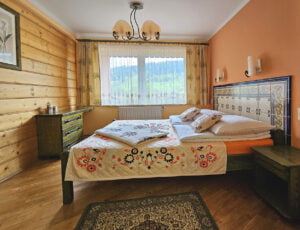 Przestronna sypialnia z łóżkiem małżeńskim, dużym oknem z widokiem na Skrzyczne, boazerią na ścianach, parkietem na podłodze, szafą oraz komodą.