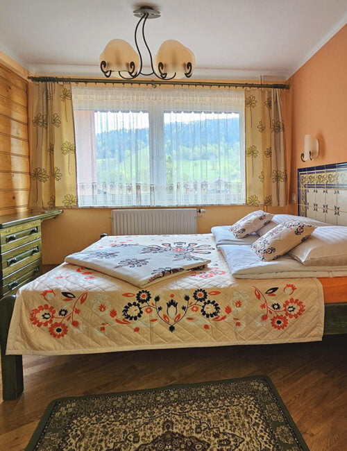 Przestronna sypialnia z łóżkiem małżeńskim, dużym oknem z widokiem na Skrzyczne, boazerią na ścianach, parkietem na podłodze, szafą oraz komodą.