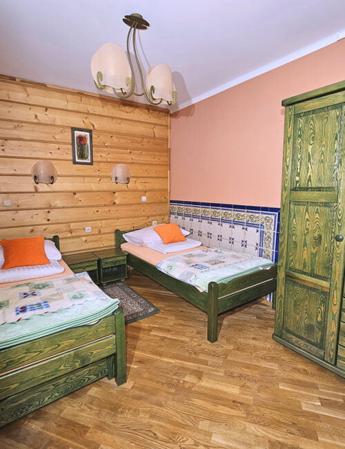 Przytulna sypialnia utrzymana w ciepłych barwach, z dwoma drewnianymi łóżkami pojedynczymi, oknem z widokiem na ogród, boazerią oraz szafą.