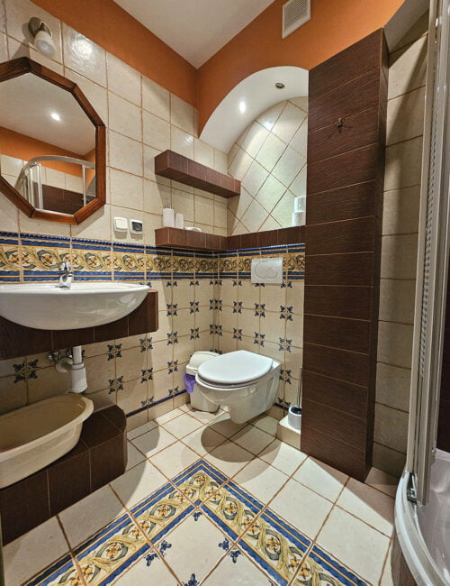 Stylowa łazienka utrzymana w jasno brązowych barwach, wyposażona w kabinę prysznicową, umywalkę, toaletę, suszarkę, grzejnik oraz ręczniki.