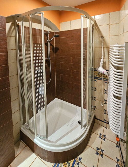 Stylowa łazienka utrzymana w jasno brązowych barwach, wyposażona w kabinę prysznicową, umywalkę, toaletę, suszarkę, grzejnik oraz ręczniki.