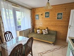 Przestronny salon z balkonem, wyposażony w kanapę, stół oraz aneks kuchenny w kremowo-oliwkowych barwach, z boazerią na ścianie.