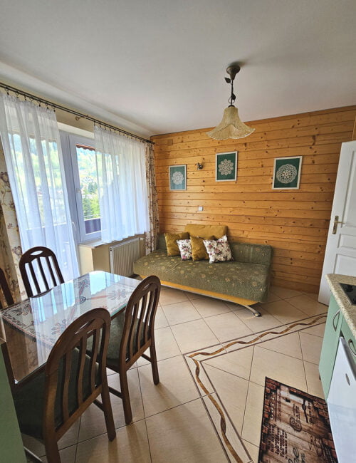 Przestronny salon z balkonem, wyposażony w kanapę, stół oraz aneks kuchenny w kremowo-oliwkowych barwach, z boazerią na ścianie.