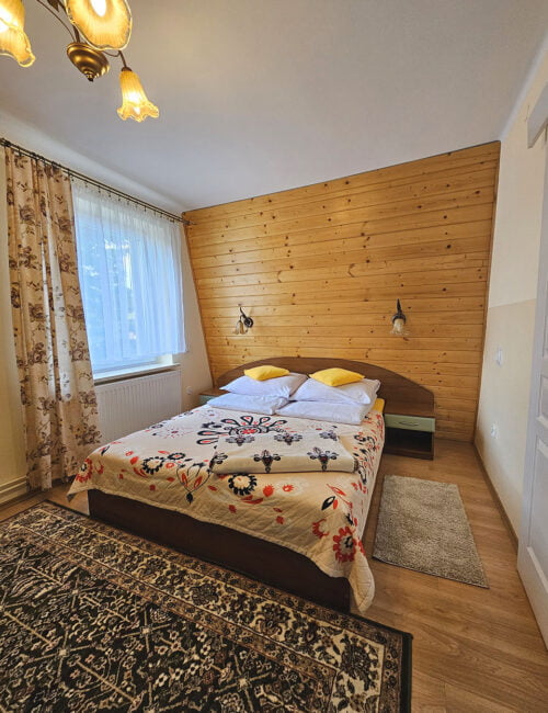 Przytulna sypialnia z łóżkiem małżeńskim, utrzymana w ciepłych barwach, z oknem, boazerią oraz wiosenną tapetą i szafą