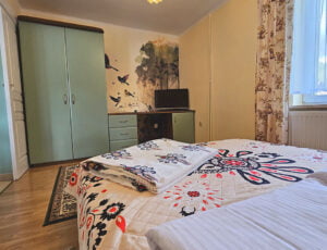 Przyjemna sypialnia urządzona w ciepłych barwach, z wiosenną tapetą dodającą świeżości. Dodatkowo, okno, boazeria oraz elegancka szafa kompleksują atmosferę tego wnętrza.