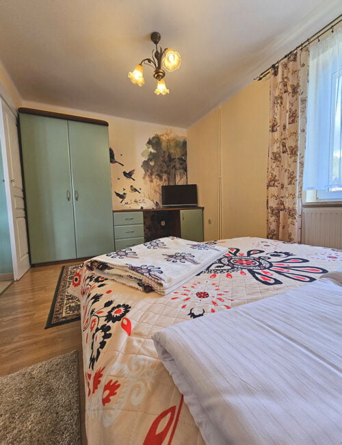 Przyjemna sypialnia urządzona w ciepłych barwach, z wiosenną tapetą dodającą świeżości. Dodatkowo, okno, boazeria oraz elegancka szafa kompleksują atmosferę tego wnętrza.