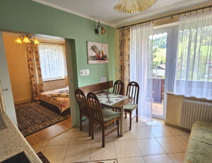 Przestronny salon z malowniczym balkonem, kanapą oraz stołem, otwarty na aneks kuchenny w kolorach kremowo-oliwkowych. Boazeria na ścianie dodaje wyjątkowego uroku temu wnętrzu