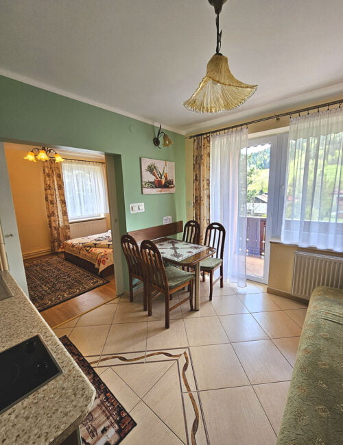 Przestronny salon z malowniczym balkonem, kanapą oraz stołem, otwarty na aneks kuchenny w kolorach kremowo-oliwkowych. Boazeria na ścianie dodaje wyjątkowego uroku temu wnętrzu