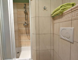 Przyjemna łazienka w kremowo-zielonych barwach, wyposażona w kabinę prysznicową, grzejnik, umywalkę, toaletę, suszarkę oraz ręczniki.