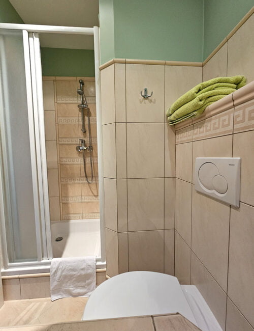Przyjemna łazienka w kremowo-zielonych barwach, wyposażona w kabinę prysznicową, grzejnik, umywalkę, toaletę, suszarkę oraz ręczniki.