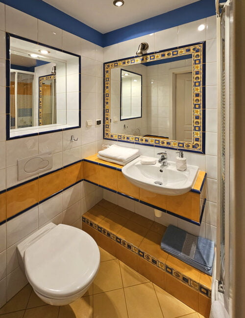 Jasna łazienka z kabina prysznicową, toaletą, umywalką, dwoma lustrami, grzejnikiem, suszarką i ręcznikami.