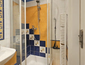 Przyjemna, jasna łazienka ożywiona delikatnymi kolorami, wyposażona w kabinę prysznicową, toaletę, umywalkę, dwie lustra, grzejnik, suszarkę i kolorowe ręczniki.