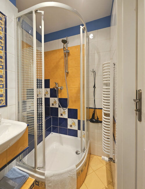 Przyjemna, jasna łazienka ożywiona delikatnymi kolorami, wyposażona w kabinę prysznicową, toaletę, umywalkę, dwie lustra, grzejnik, suszarkę i kolorowe ręczniki.