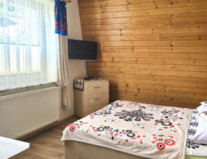 Przytulna sypialnia w kremowych tonacjach z boazerią na ścianie, dużym oknem, obrazami zimowymi, łóżkiem małżeńskim i telewizorem