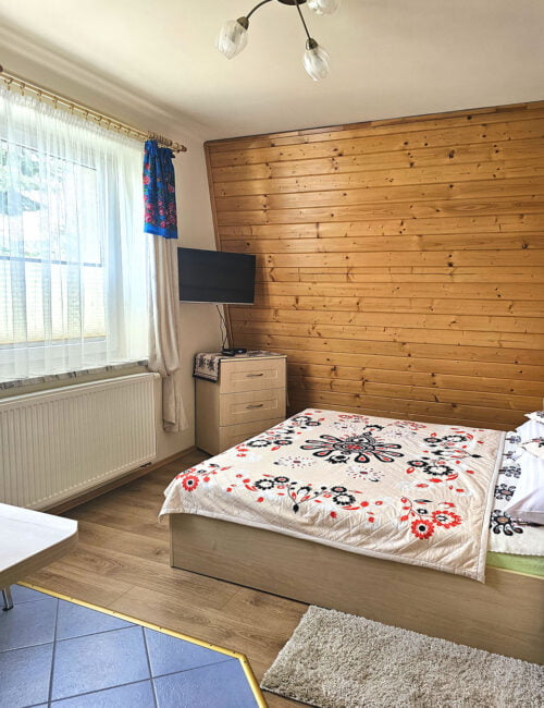 Przytulna sypialnia w kremowych tonacjach z boazerią na ścianie, dużym oknem, obrazami zimowymi, łóżkiem małżeńskim i telewizorem