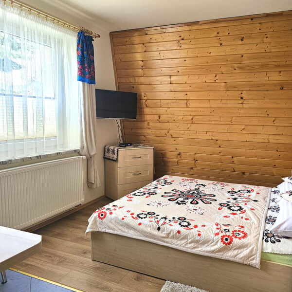 Przytulna sypialnia w kremowych tonacjach z boazerią na ścianie, dużym oknem, obrazami zimowymi, łóżkiem małżeńskim i telewizorem