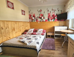 Przytulna sypialnia w kremowych tonacjach z łóżkiem małżeńskim, boazerią, tv oraz barwną tapetą. Uroku dodają góralskie dodatki.