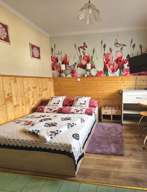 Przytulna sypialnia w kremowych tonacjach z łóżkiem małżeńskim, boazerią, tv oraz barwną tapetą. Uroku dodają góralskie dodatki.