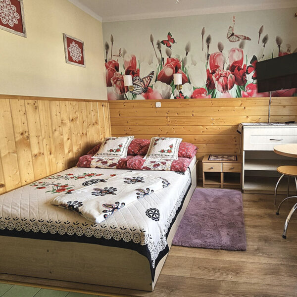 Przytulna sypialnia w kremowych tonacjach z łóżkiem małżeńskim, boazerią, tv oraz barwną tapetą. Uroku dodają góralskie dodatki.