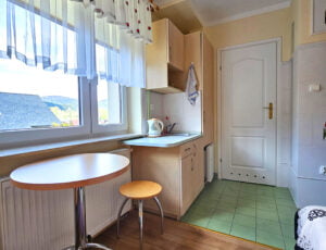 Aneks kuchenny z zlewem, lodówką i czajnikiem, duże okno ozdobione uroczymi zasłonkami, oraz stolik dla dwóch osób