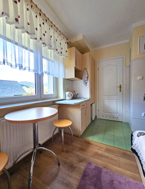 Aneks kuchenny z zlewem, lodówką i czajnikiem, duże okno ozdobione uroczymi zasłonkami, oraz stolik dla dwóch osób