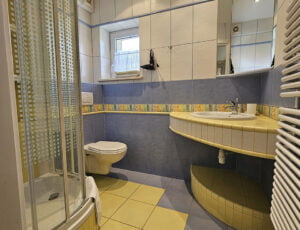 Łazienka w biało-żółto-niebieskich barwach z toaletą, umywalką, kabiną prysznicową, suszarką i kolorowymi ręcznikami.