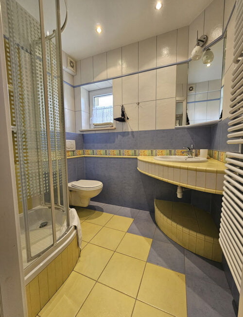 Łazienka w biało-żółto-niebieskich barwach z toaletą, umywalką, kabiną prysznicową, suszarką i kolorowymi ręcznikami.