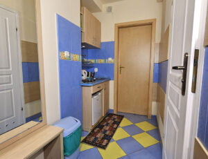 Aneks kuchenny w kremowo-niebieskich barwach z zlewem, płytą grzewczą, czajnikiem, lodówka oraz lustrem na ścianie.