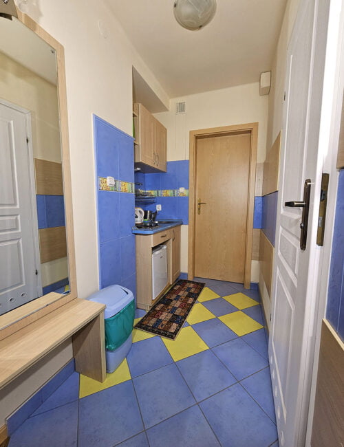 Aneks kuchenny w kremowo-niebieskich barwach z zlewem, płytą grzewczą, czajnikiem, lodówka oraz lustrem na ścianie.