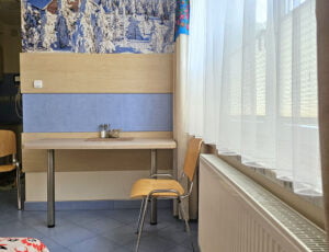 Sypialnia z dużym oknem i stolikiem dla dwóch osób, zdobiona obrazem przedstawiającym zimową scenerię ze schroniskiem na Skrzycznem.